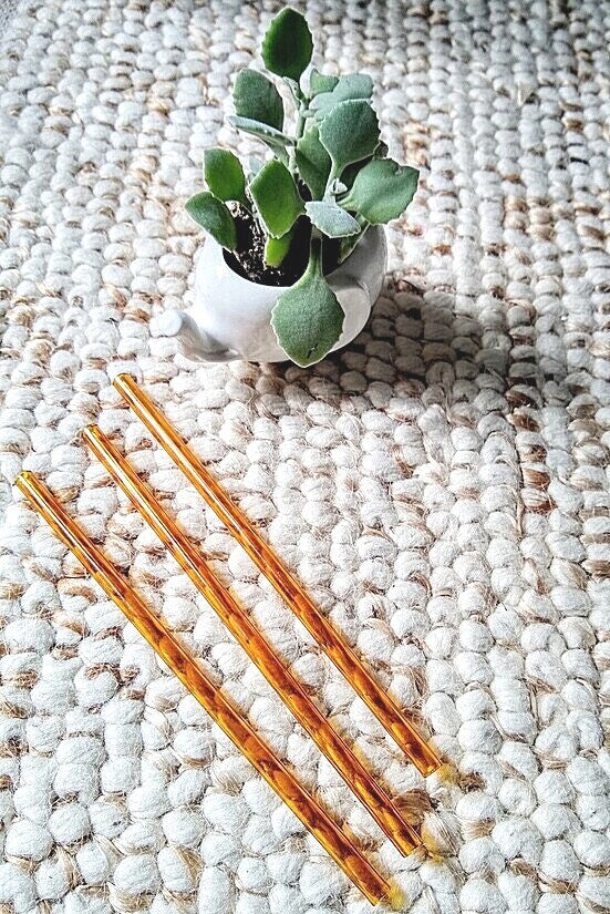 Photo of three ToMA glass straws in artisanal amber on textured carpet with small white porcelain elephant planter with succulent. 